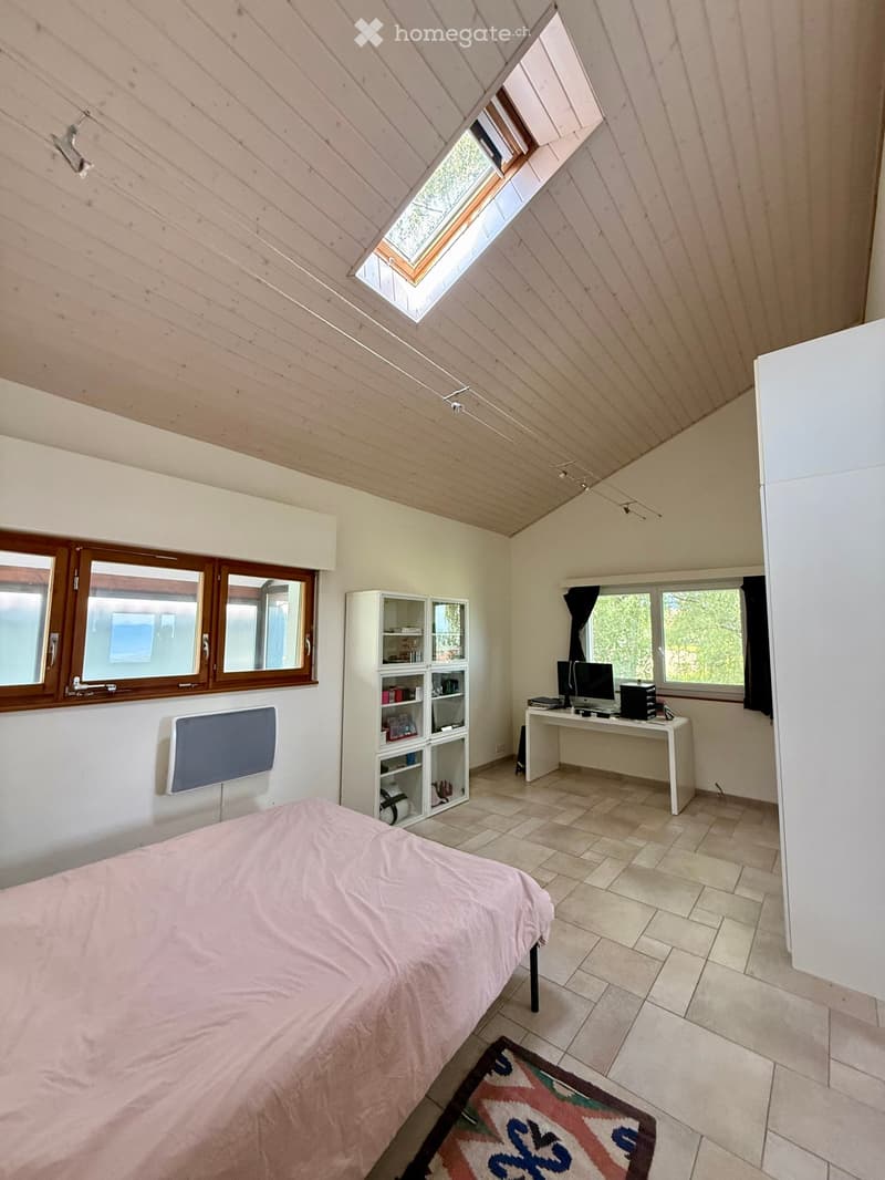 Bright bedroom with a bed, desk, computer, shelf, and mirror. The room features wooden slatted ceilings and large windows, including a skylight.