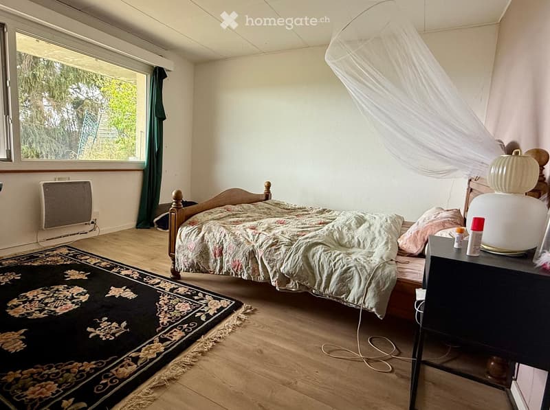 Cozy bedroom with wooden floor, large window with green curtain, bed with floral duvet, nightstand with lamp, radiator, and patterned rug.