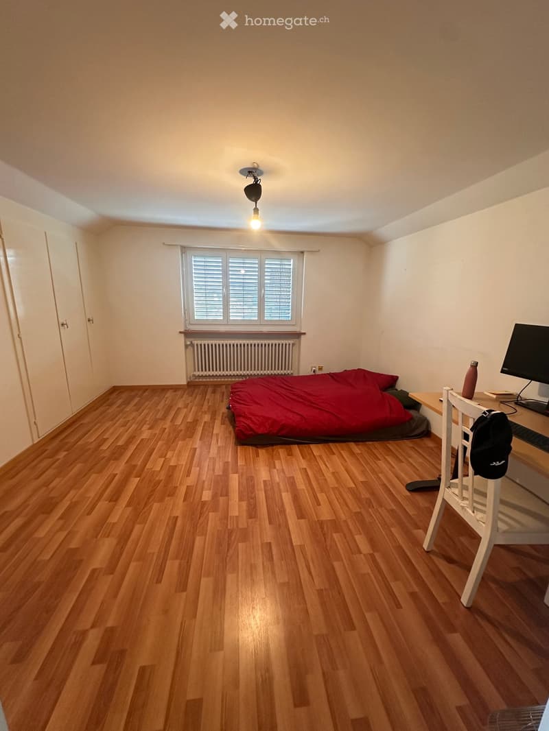 A small, furnished bedroom with wooden flooring, a bed with red bedding, a desk with a chair, a screen, and a backpack. The room has a window with blinds and a radiator below it.