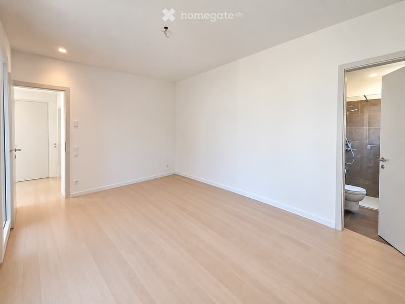 Empty room with light wooden flooring, white walls, and a partially visible bathroom with a toilet and shower stall.