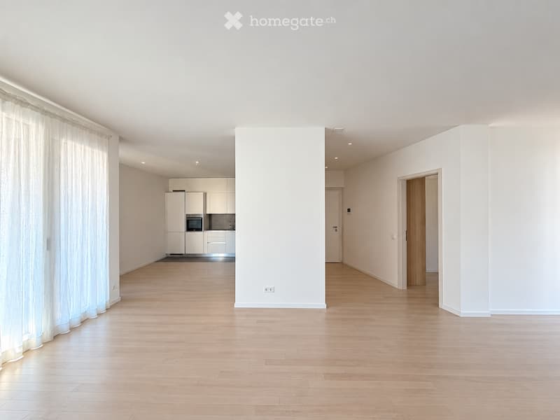 Empty room with light wooden flooring, white walls, and a large window with sheer curtains. The room is connected to a small kitchen area with white cabinets and a built-in oven.
