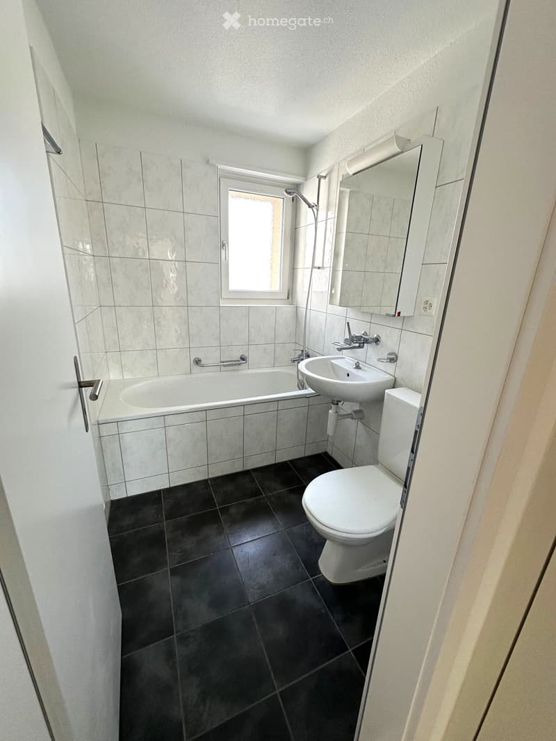Modern bathroom with a bathtub, sink, toilet, and mirror. Features dark tiled flooring and light grey tiled walls.