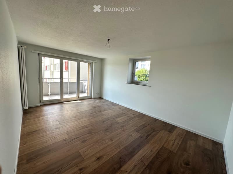 Empty room with wooden flooring, white walls, large sliding glass doors leading to a balcony, and a small window with a view of greenery.
