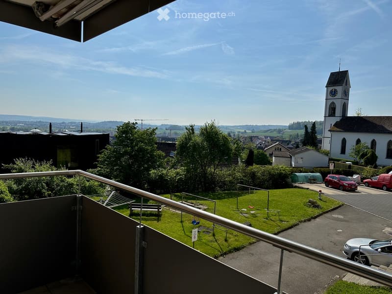 Balcony overlooking a green lawn, playground, and a town with a church tower in the background.