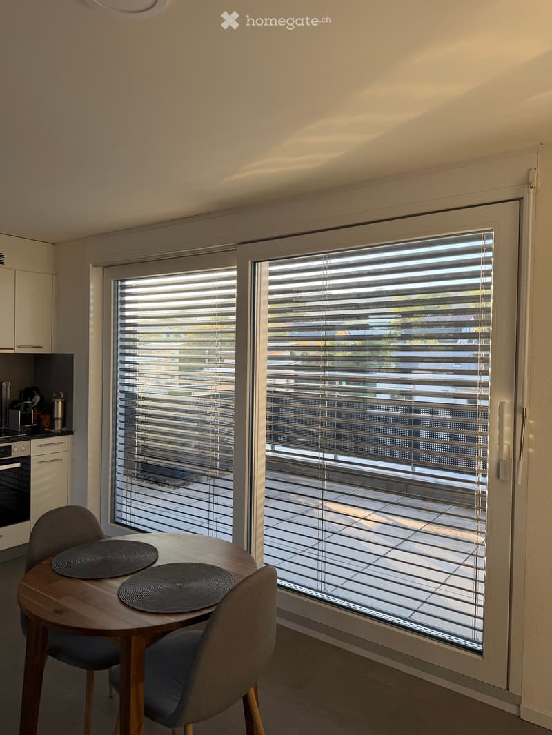 A modern dining area with a round wooden table, two chairs, and a view of a balcony through large windows with blinds. The kitchen area includes an oven and a small cabinet.