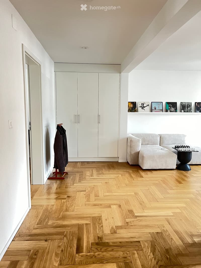 Modern living room with herringbone wooden flooring, white walls, a grey sectional sofa, a small black table with a chessboard, a coat rack with a jacket, and built-in white cabinets with framed artwork on the wall.