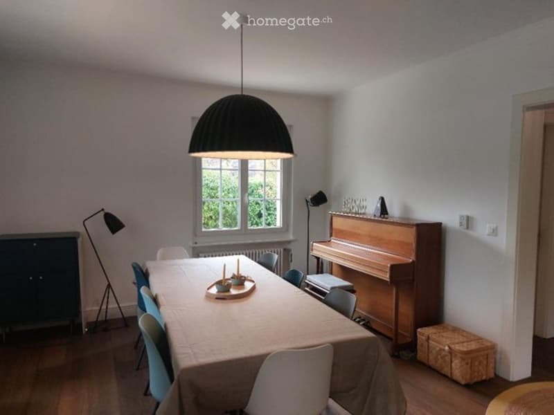 Dining room with a long wooden table covered in a beige tablecloth, surrounded by blue and white chairs. A grand piano is positioned near the window, accompanied by a small stool. Two floor lamps and a table lamp provide lighting. A dark wooden cabinet is