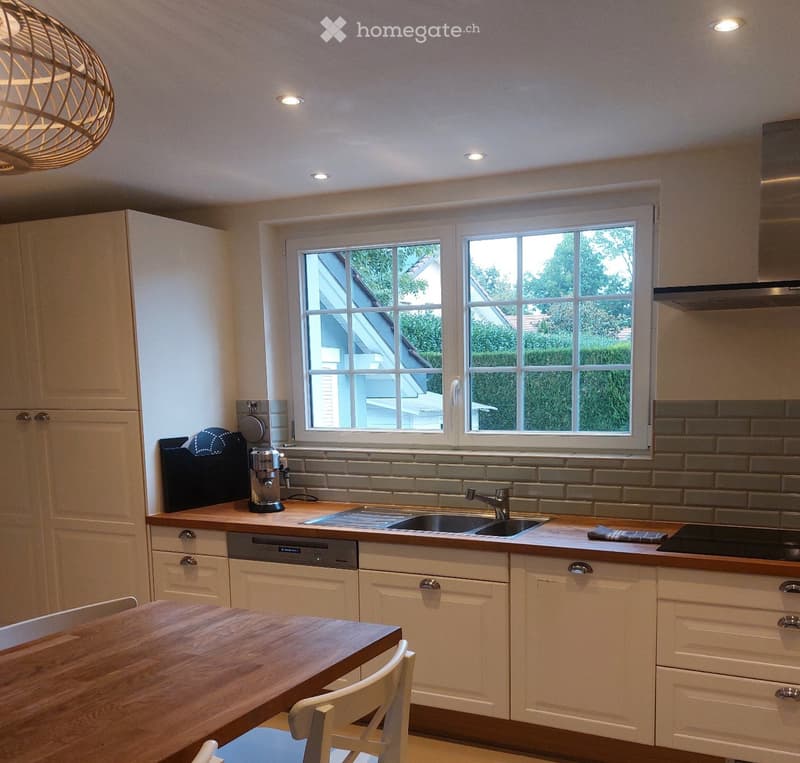 Modern kitchen with light-colored cabinets, wooden countertops, a sink, a dishwasher, a coffee maker, and a dining area with a wooden table and chairs.