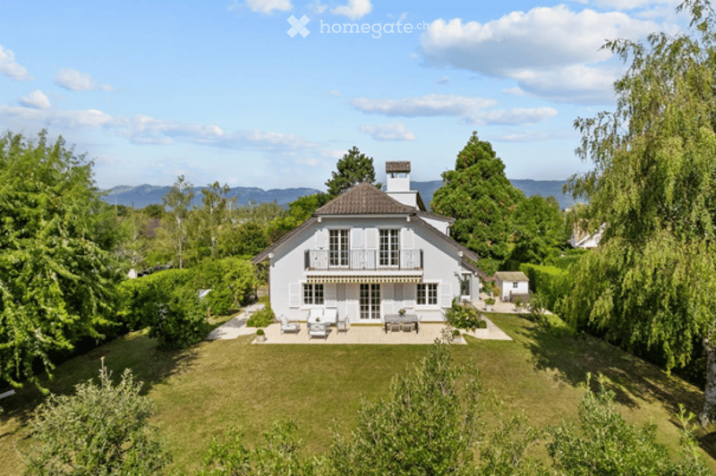 A two-story white house with a tiled roof, surrounded by a well-maintained garden and trees. The house features a balcony and outdoor seating area with chairs and a table.