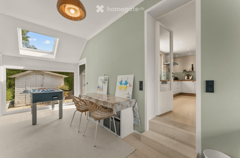 The room is a modern, open-concept space with a dining area and a lounge area. It features a round wooden table with four wicker chairs, a futon, and a small kitchen area with a fridge, cupboard, and stool. There is a large window with a view of the outdo