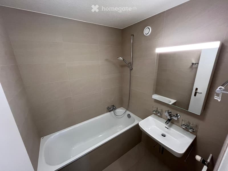 Modern bathroom with a bathtub, sink, mirror, and showerhead. The walls are tiled in a neutral color, and the space is well-lit with a built-in light above the mirror.