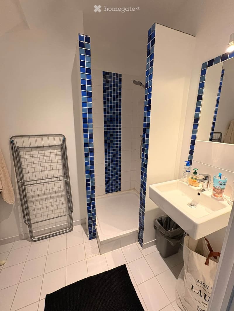 Modern bathroom with white tiles, a bathtub, a sink with a mirror, a towel rack, and a trash bin. Blue mosaic tiles decorate the shower area.