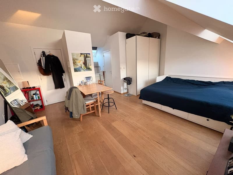 A small, well-lit bedroom with a bed, a small table with chairs, a cabinet, and a sofa. The room features wooden flooring and has a door leading to another area.