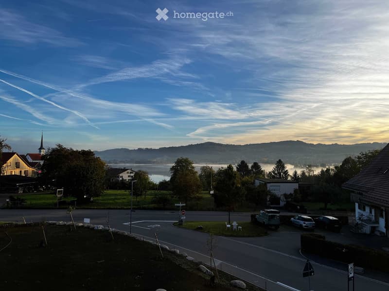 A peaceful lakeside village scene with houses, trees, a church spire, and a calm lake under a partly cloudy sky.