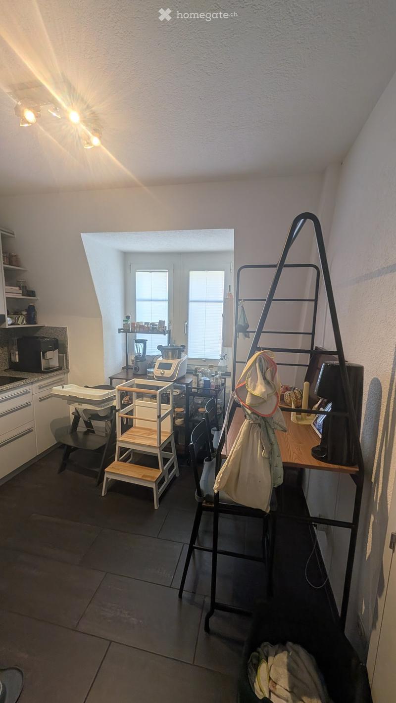 A small, well-lit room with a baby chair, a small wooden stool, a chair, a table, a cupboard, a blender, a coffee maker, and a lamp. The room has a slanted ceiling and a window with blinds.