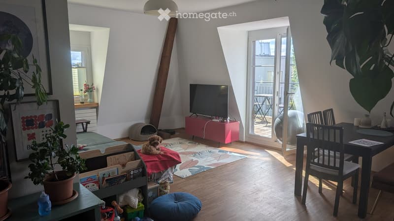 Bright living room with wooden flooring, large windows, and a balcony. Features include a sofa, cat bed, television, dining table, chairs, and various decorative items.