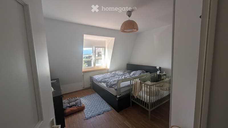 Cozy bedroom with wooden flooring, a double bed, a baby crib, a small rug, and a dog bed. The room features a large window with a view and a hanging woven lamp.