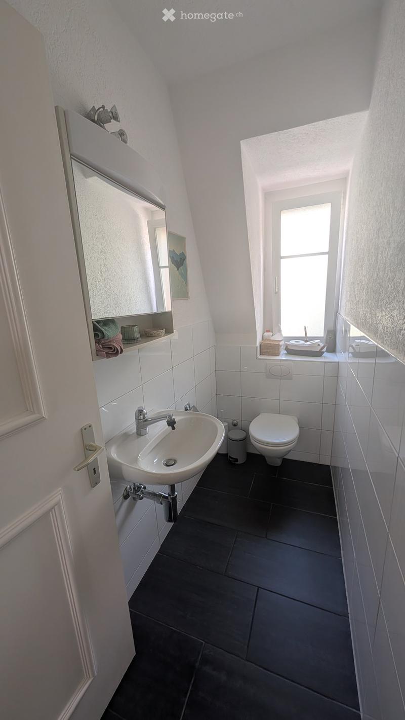 Modern bathroom with a white sink, chrome faucet, toilet, and a mirror above the sink. The room features dark tiled flooring and white tiled walls, with a window providing natural light.