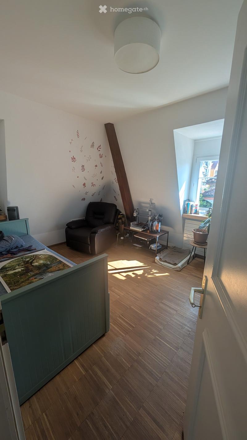 Cozy attic bedroom with wooden flooring, slanted ceilings, and large windows. Features a bed, armchair, small table, and bookshelf.