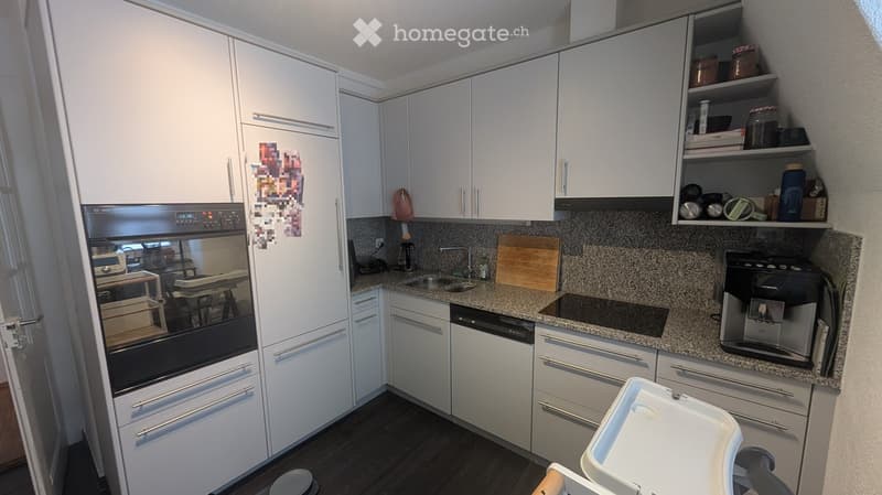 Modern kitchen with white cabinets, stainless steel appliances, granite countertops, and a built-in coffee machine. Shelves hold various items, and a baby seat is visible in the foreground.