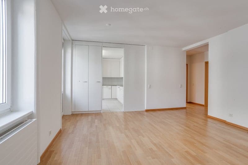 Empty room with light wooden flooring, white walls, large built-in closet, and a view into an adjacent kitchen area.