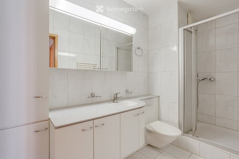 Modern bathroom with white tiles, a large mirror above the sink, a toilet, and a glass-enclosed shower. The sink has a chrome faucet and is set into a white countertop with cabinets below.