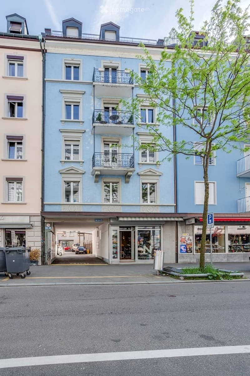 Multi-story building with light blue facade, white trim, multiple windows, balconies, and a ground-floor garage entrance.