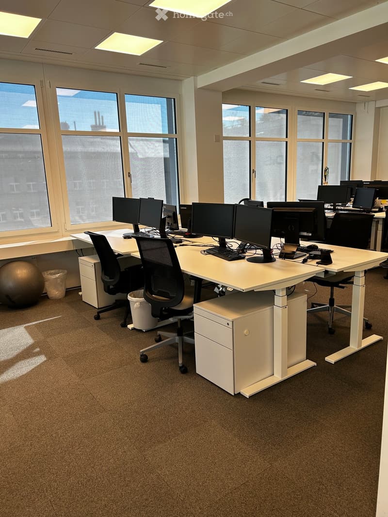 Modern office space with large windows, desks, chairs, multiple computer monitors, keyboards, mice, and a server rack. A large exercise ball and trash cans are also visible.