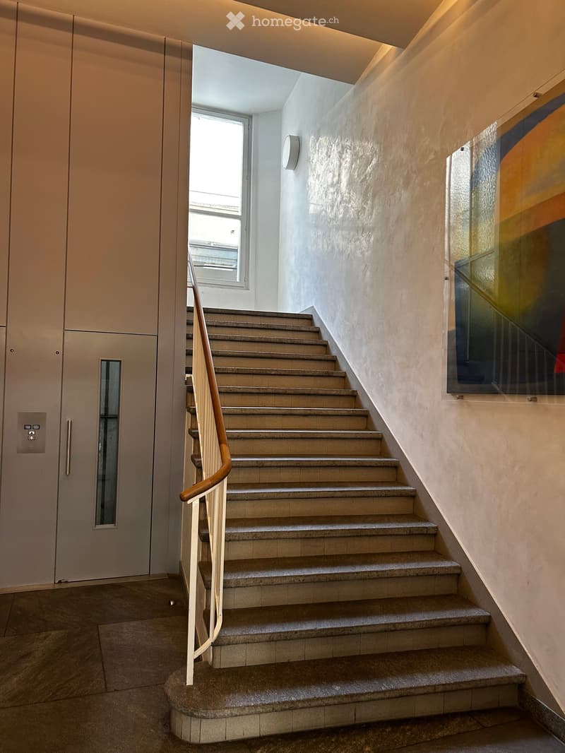 Modern staircase with beige stone steps, wooden handrail, and a wall-mounted light fixture. The hallway features a textured wall and a glass panel on one side.