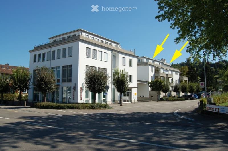 Modern multi-story building with white facade, large windows, and a flat roof, surrounded by trees and parked cars.