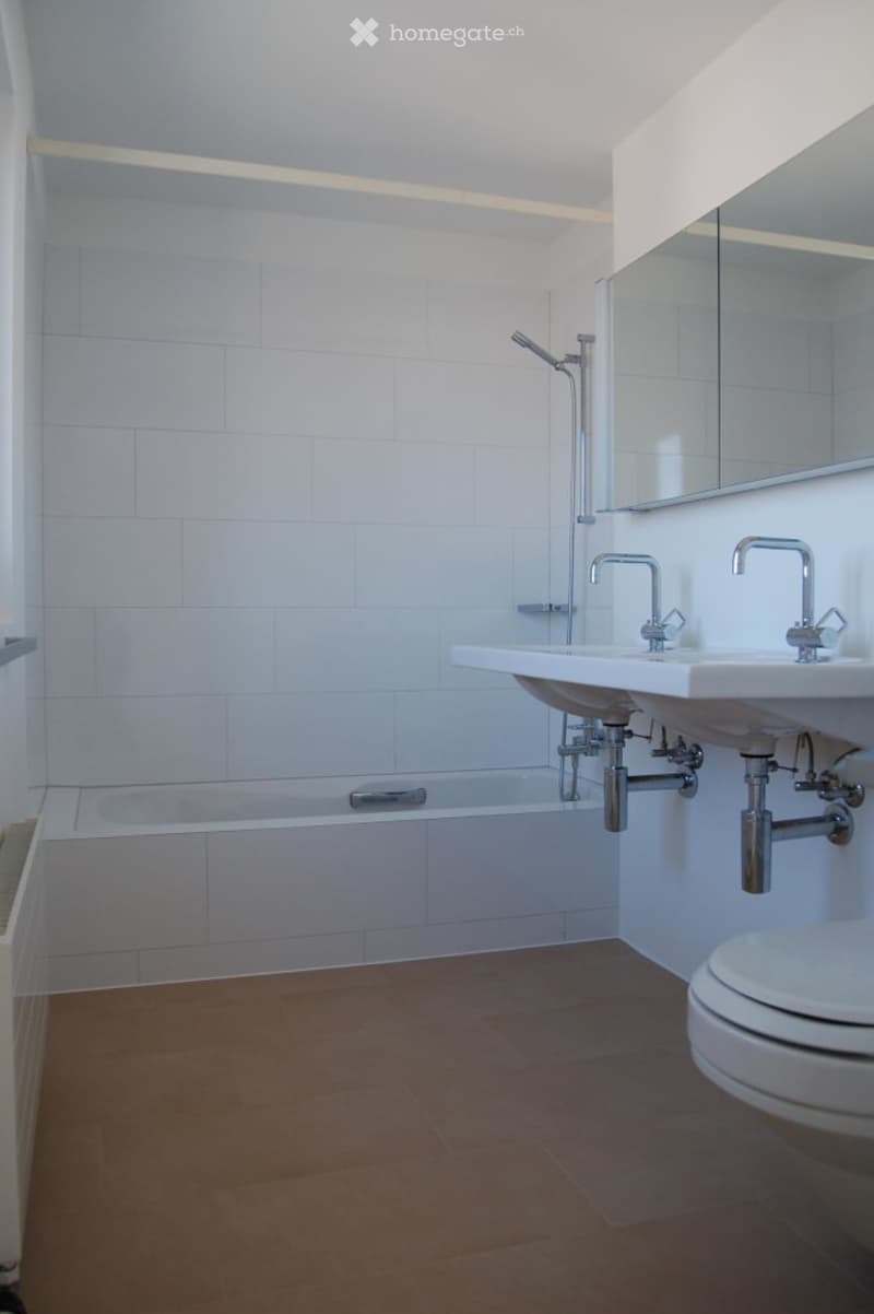 Modern bathroom with white tiled walls, a bathtub, a double sink, a toilet, and a large mirror above the sink.