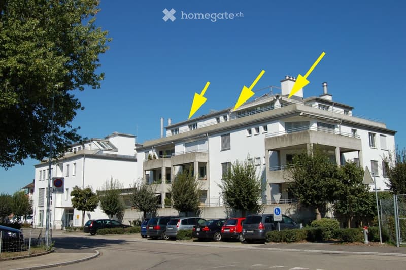 Multi-story residential building with balconies, white facade, and several parked cars in front.