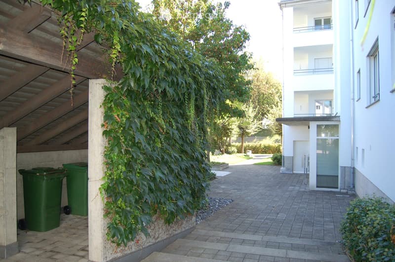 A paved outdoor area with a covered entrance, green trash bins, and a building with a balcony and greenery in the background.