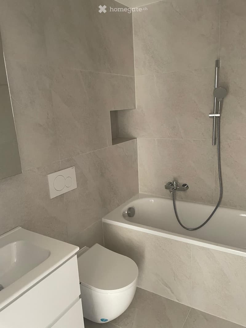 Modern bathroom with a bathtub, toilet, and showerhead. The walls are tiled in a light beige color, and the floor is also tiled in a matching shade. There is a small shelf built into the wall above the bathtub.