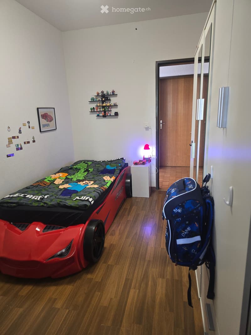 A child's bedroom with a car-shaped bed, a backpack hanging on the closet, a small shelf with toys, a lamp, and a picture frame on the wall.