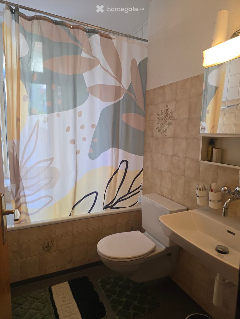 Bathroom with beige tiles, a toilet, a sink, a bathtub with a colorful shower curtain, and a mirror above the sink. There are two rugs on the floor and a toothbrush holder on the sink counter.