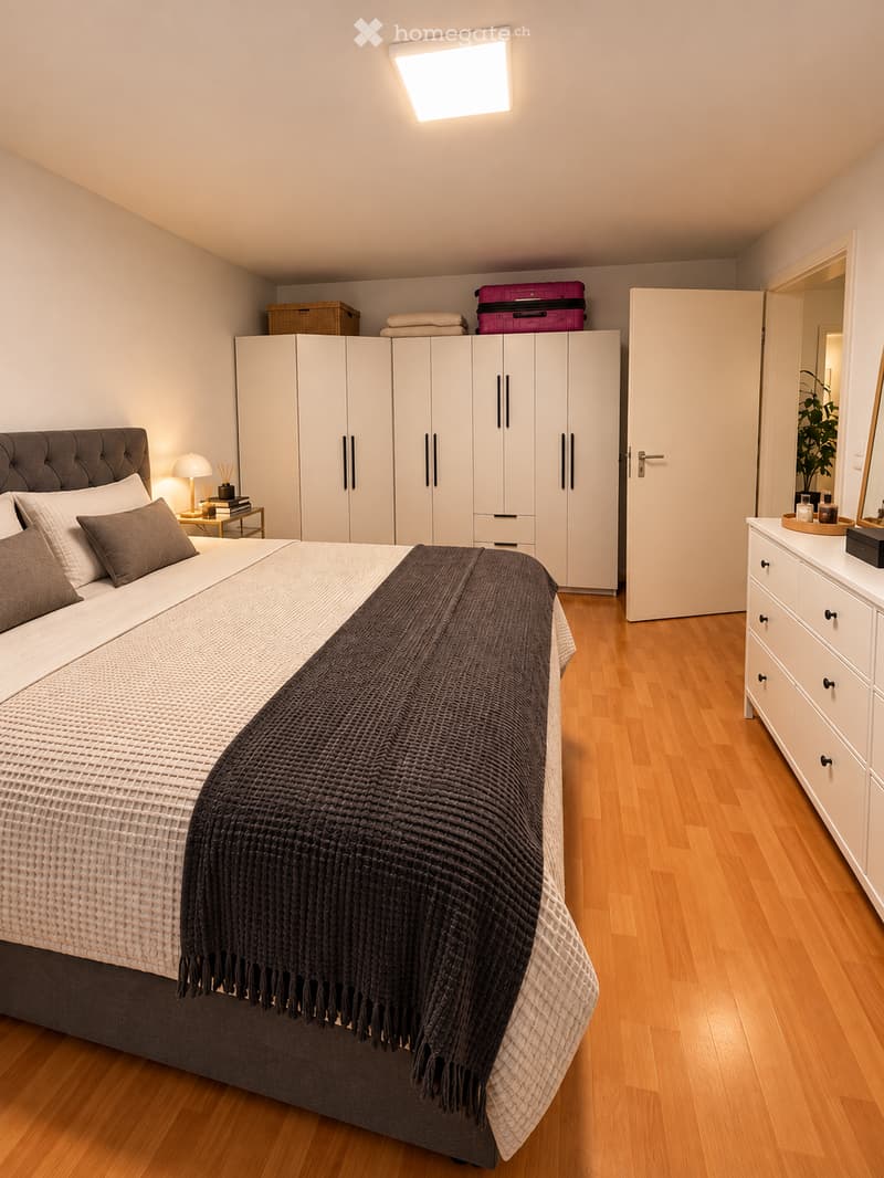 Modern bedroom with a large bed, bedside table, wardrobe, and a small plant on the dresser. The room features wooden flooring and white walls.