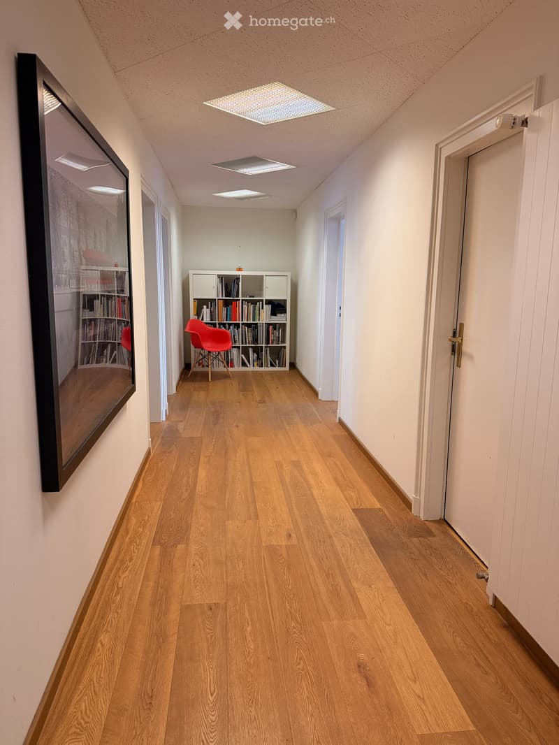 A long, narrow hallway with light wooden flooring, white walls, and recessed ceiling lighting. A small bookshelf with books and a red chair is visible in the distance, along with a large mirror on the left wall.