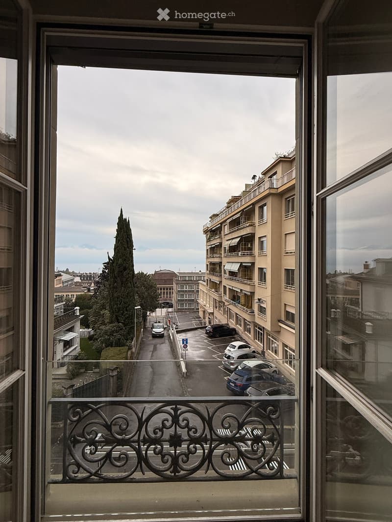 Balcony with ornate metal railing overlooking a street lined with parked cars and residential buildings.