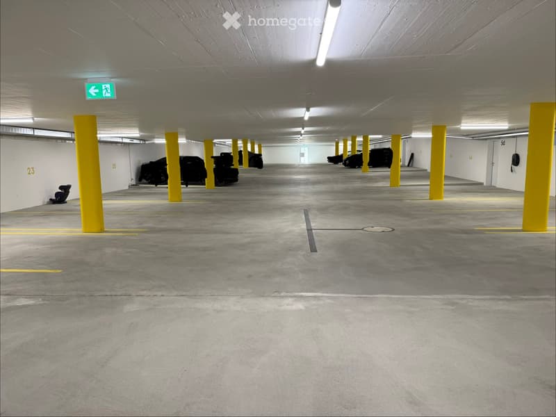 Underground parking area with concrete floors, yellow pillars, and several parked cars. The area is well-lit with fluorescent lighting and marked parking spaces.