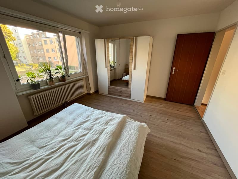 Bright bedroom with wooden flooring, large window with potted plants on the sill, a single bed with white bedding, a white wardrobe with mirrored doors, and a radiator below the window.