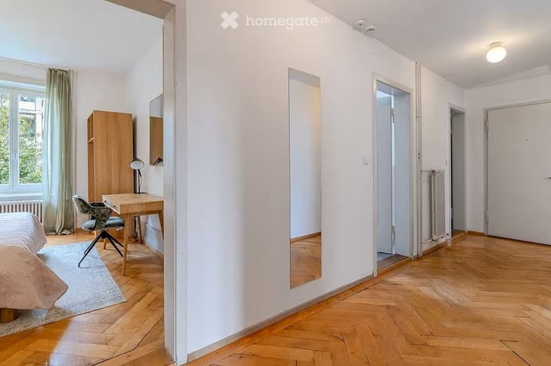 A furnished room with a bed, desk, chair, wardrobe, and mirror. The room features wooden flooring and white walls.