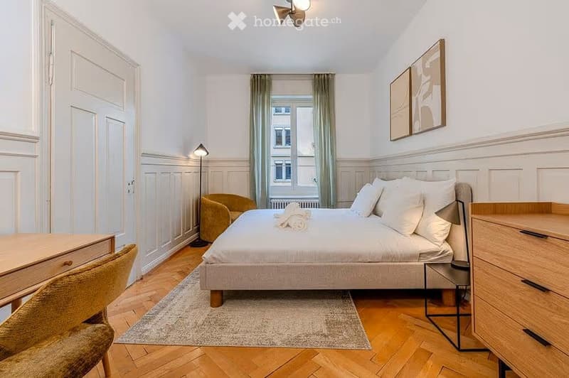 Cozy bedroom with a double bed, nightstand, chair, small table, and artwork on the wall. Wooden flooring and large window with curtains.