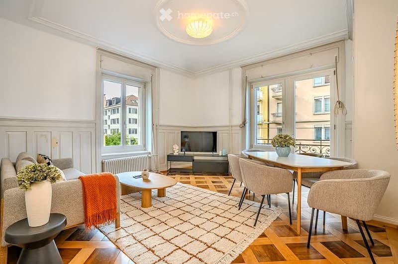 Bright living room with large windows, wooden flooring, a sofa with a throw, coffee table, dining table with chairs, and a television set.