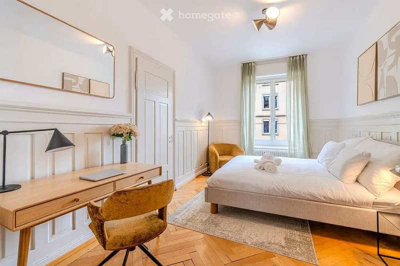 Elegant bedroom with a large bed, wooden desk, armchair, and decorative vase with flowers. The room features light-colored walls, wooden flooring, and large windows with curtains.