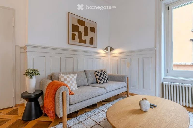 Modern living room with grey sofa, wooden coffee table, black stool, decorative vases, and artwork on the wall.
