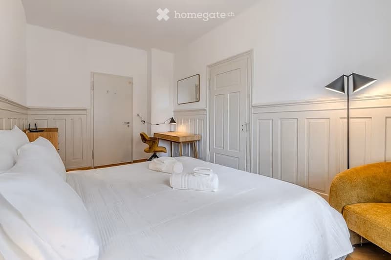 Elegant bedroom with a large white bed, neatly arranged towels, a yellow armchair, a wooden desk with a chair, a mirror, and a floor lamp.