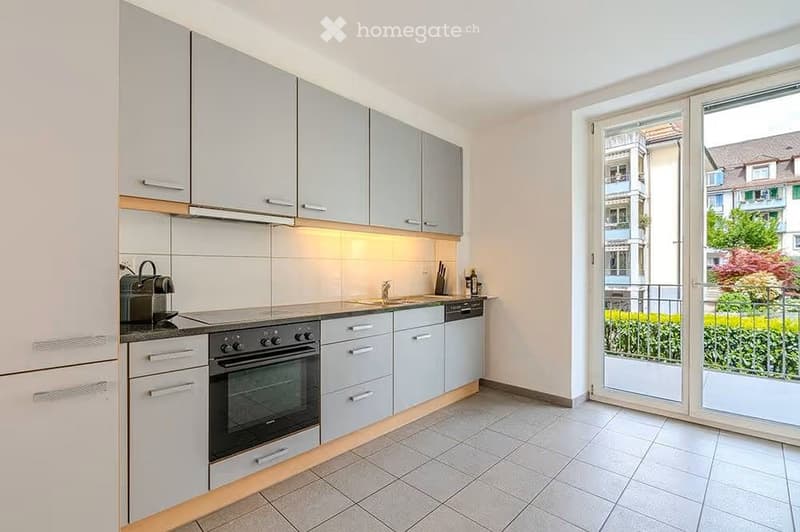 Modern kitchen with light grey cabinets, stainless steel appliances, and a large window opening to a balcony.