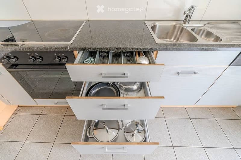 Modern kitchen with white cabinets, stainless steel sink, and open drawers revealing neatly organized pots and pans.