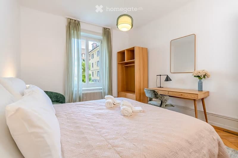 Cozy bedroom with a large bed, wooden desk, chair, bookshelf, and a mirror. The room features a window with curtains and a view of the city.